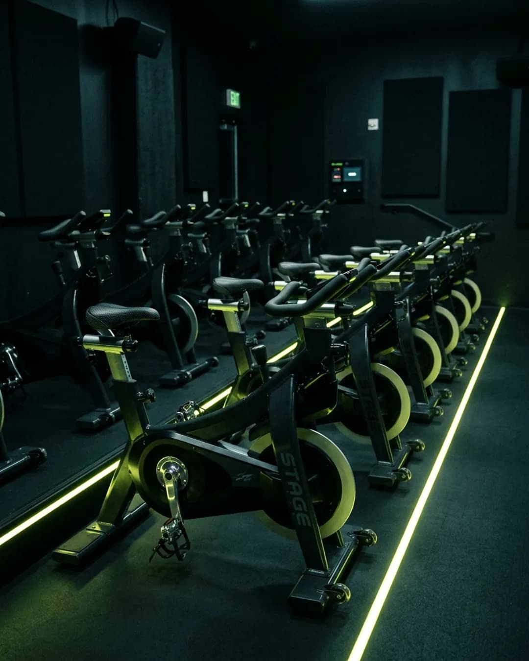 Studio de cycling en gradins, vélos noirs alignés et soulignés d'un filet de lumière vert acide.